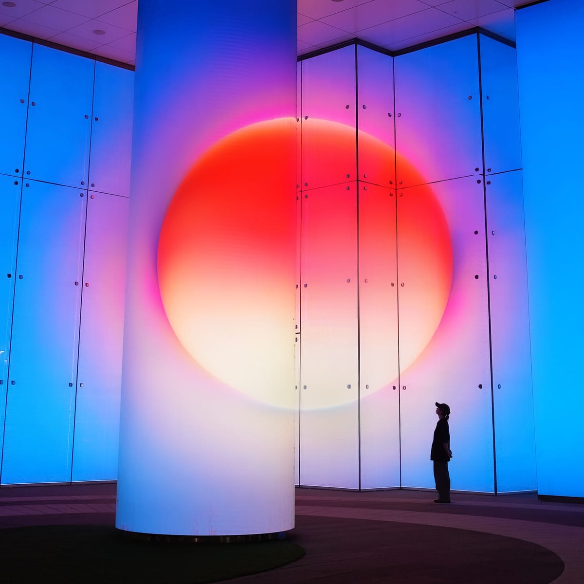 A huge red-orange sun spans a white column and blue-lit glass walls, with a visitor standing at the right for scale.