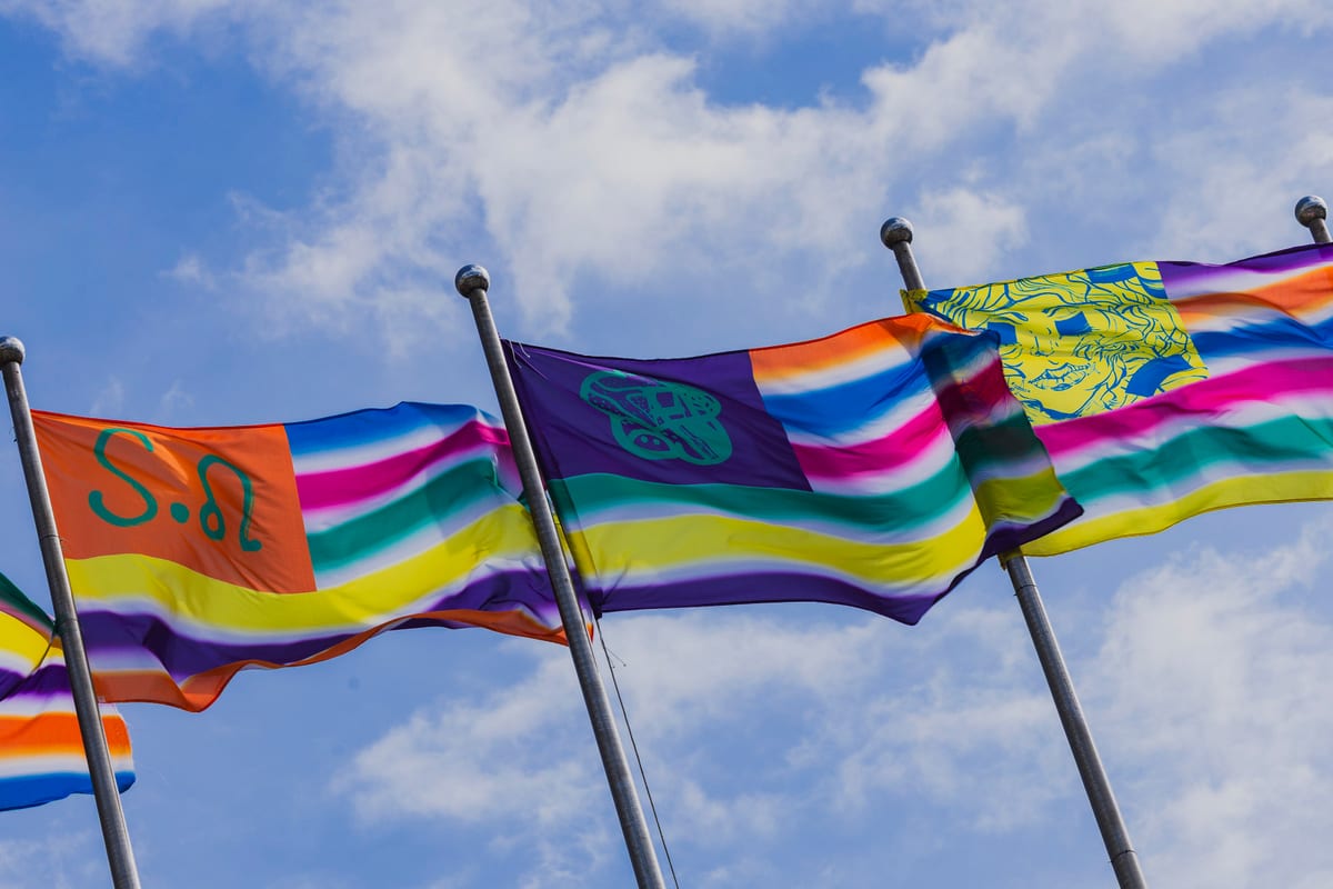 Colourful striped flags on poles around the Expo Dome carry lettering, graphic symbols and a yellow profile against the blue sky.