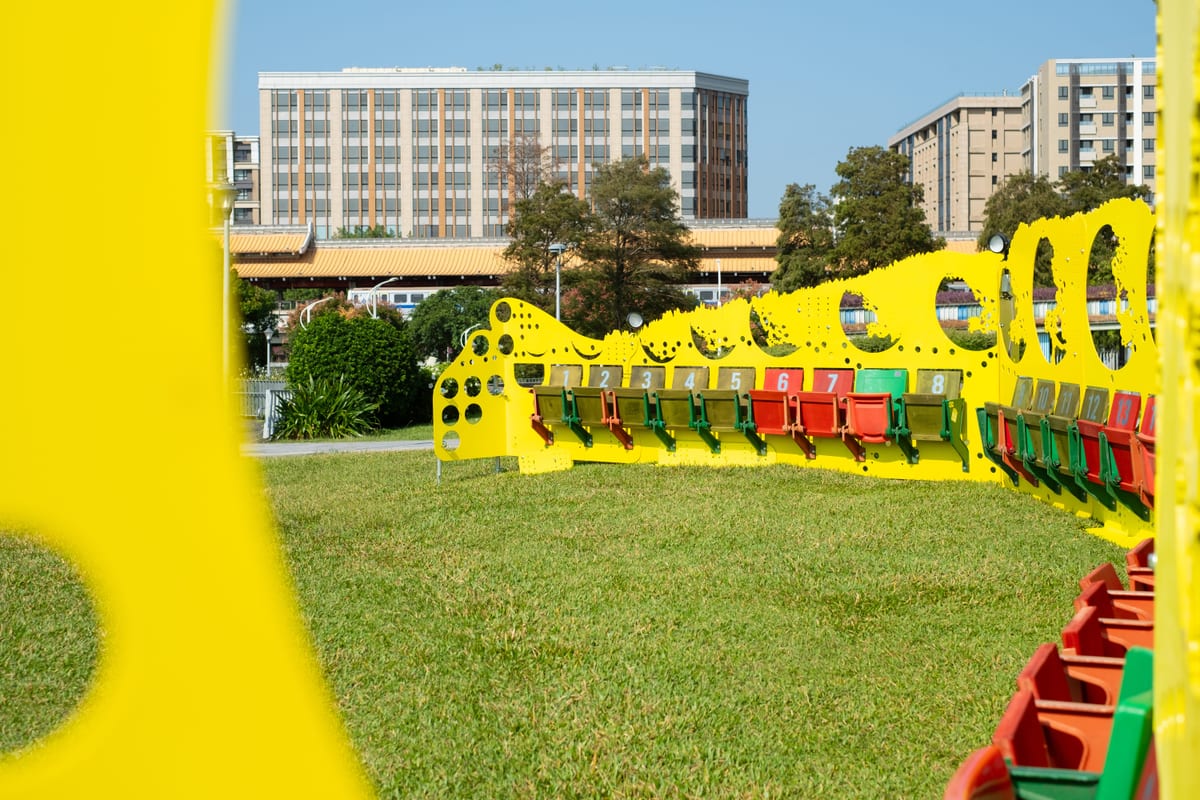 Numbered red, green and yellow folding seats line a bright yellow steel enclosure on the grass, with circular cut-outs framing buildings in Yuanshan.