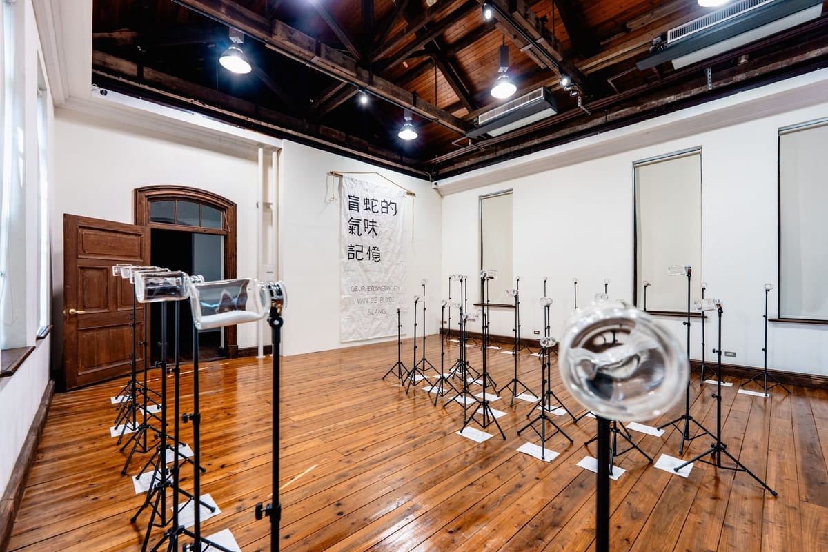Clear glass bottles lie horizontally on black tripod stands in a gallery with timber flooring and exposed rafters; a cloth at the back bears the work's title.