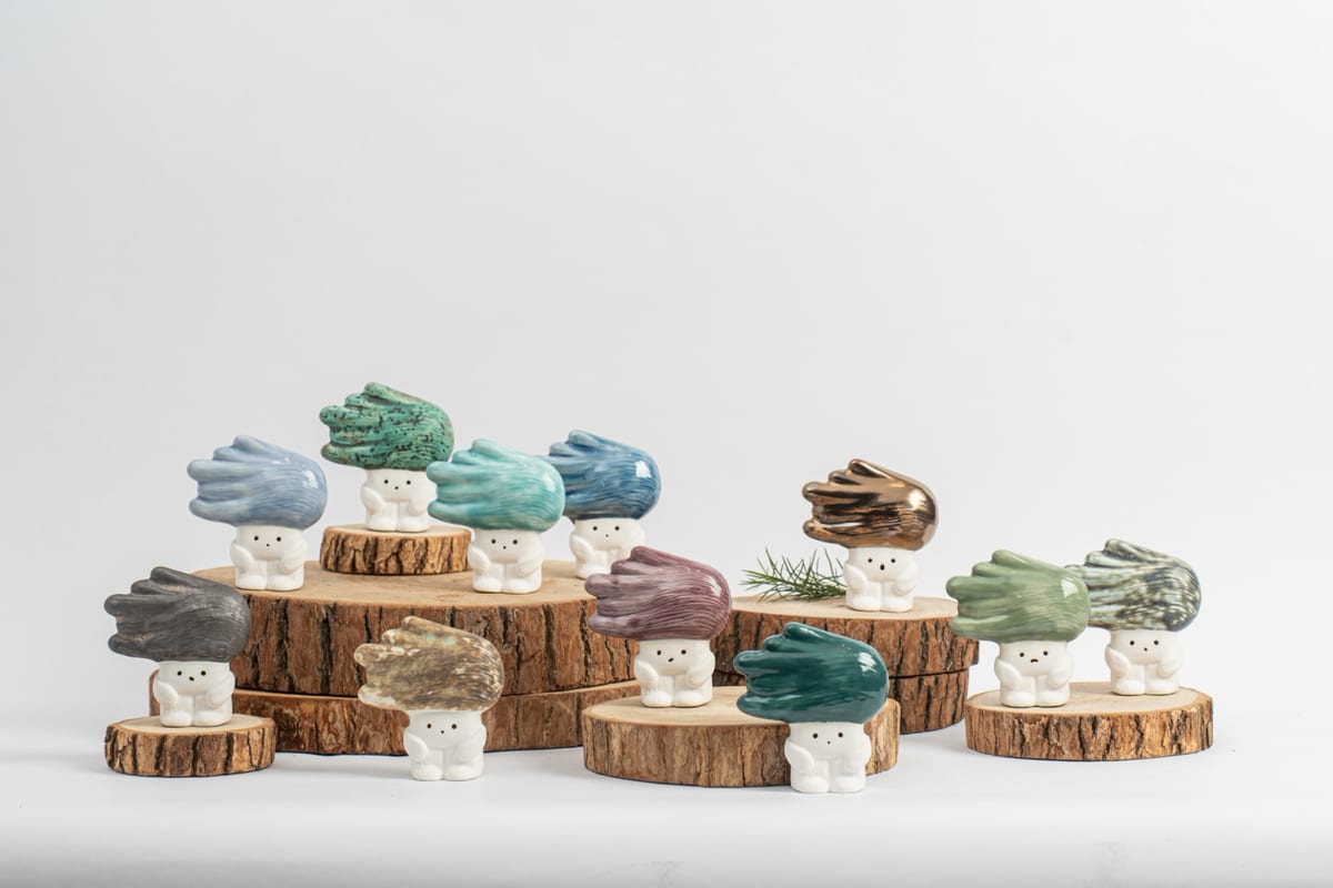 Small white ceramic tree children sit on wooden slices, their sideways crowns glazed blue, green, violet, grey and metallic brown.