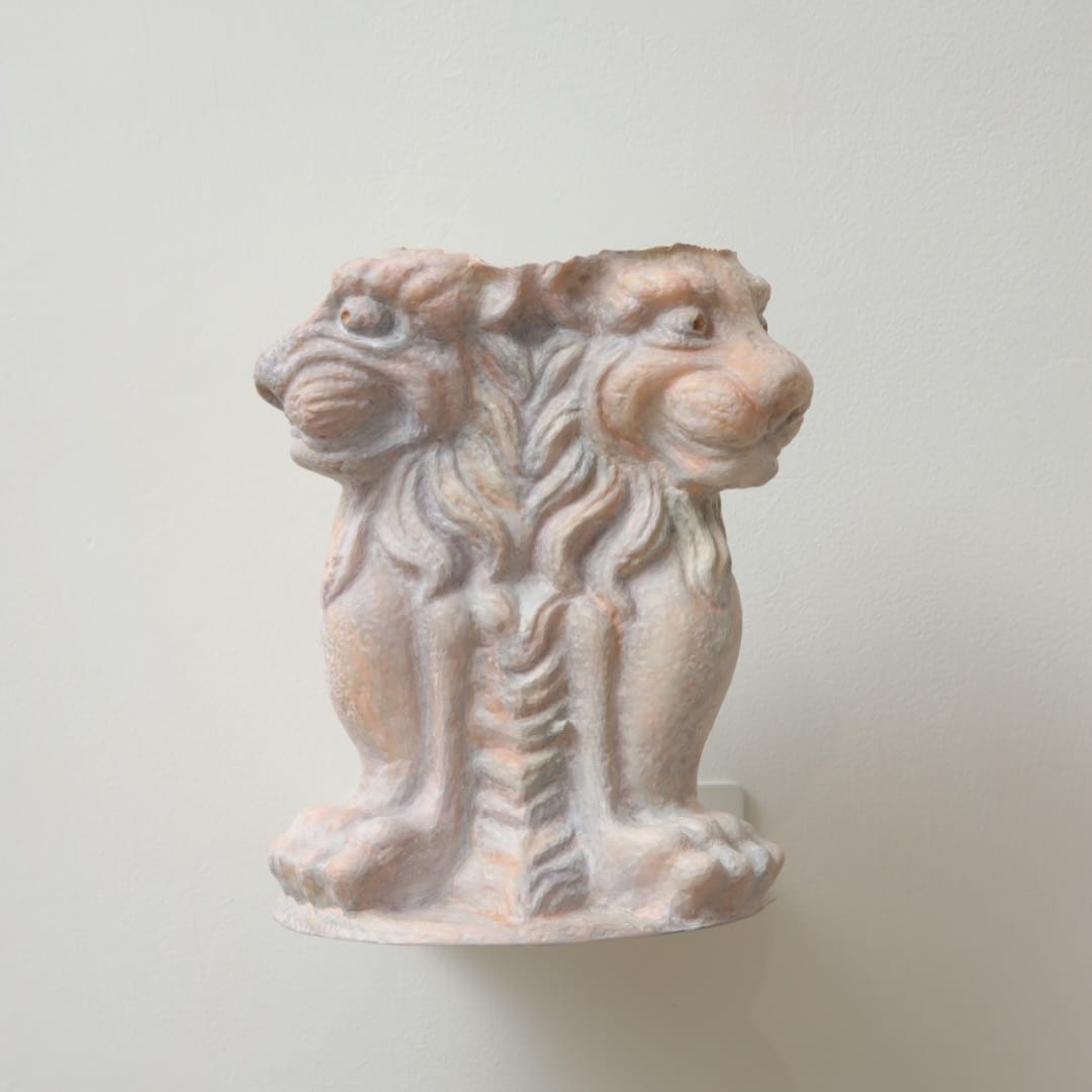 A small sculpture roughly seventeen centimetres high; a pink, gray and orange body carries a lion's face at each end, its surface covered in fine ridges and mineral flecks.