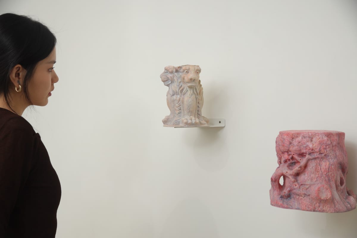 A visitor stands at a white wall looking at two small sculptures on narrow shelves, with granular brown material and a larger installation covering the floor below.