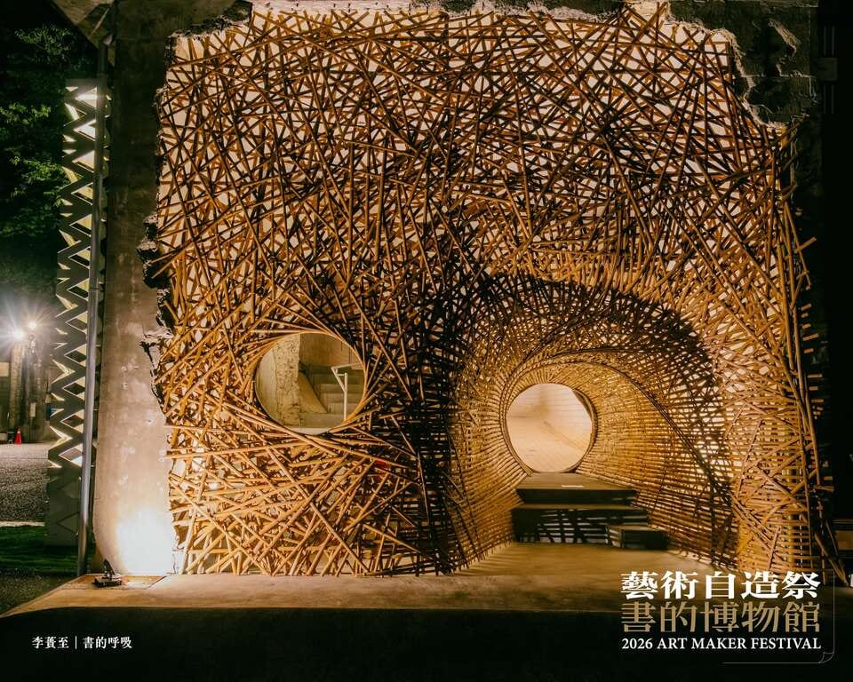 Under night lighting, densely woven bamboo fills a broken opening in an old factory wall, curling on the right into a round tunnel a visitor can walk into.