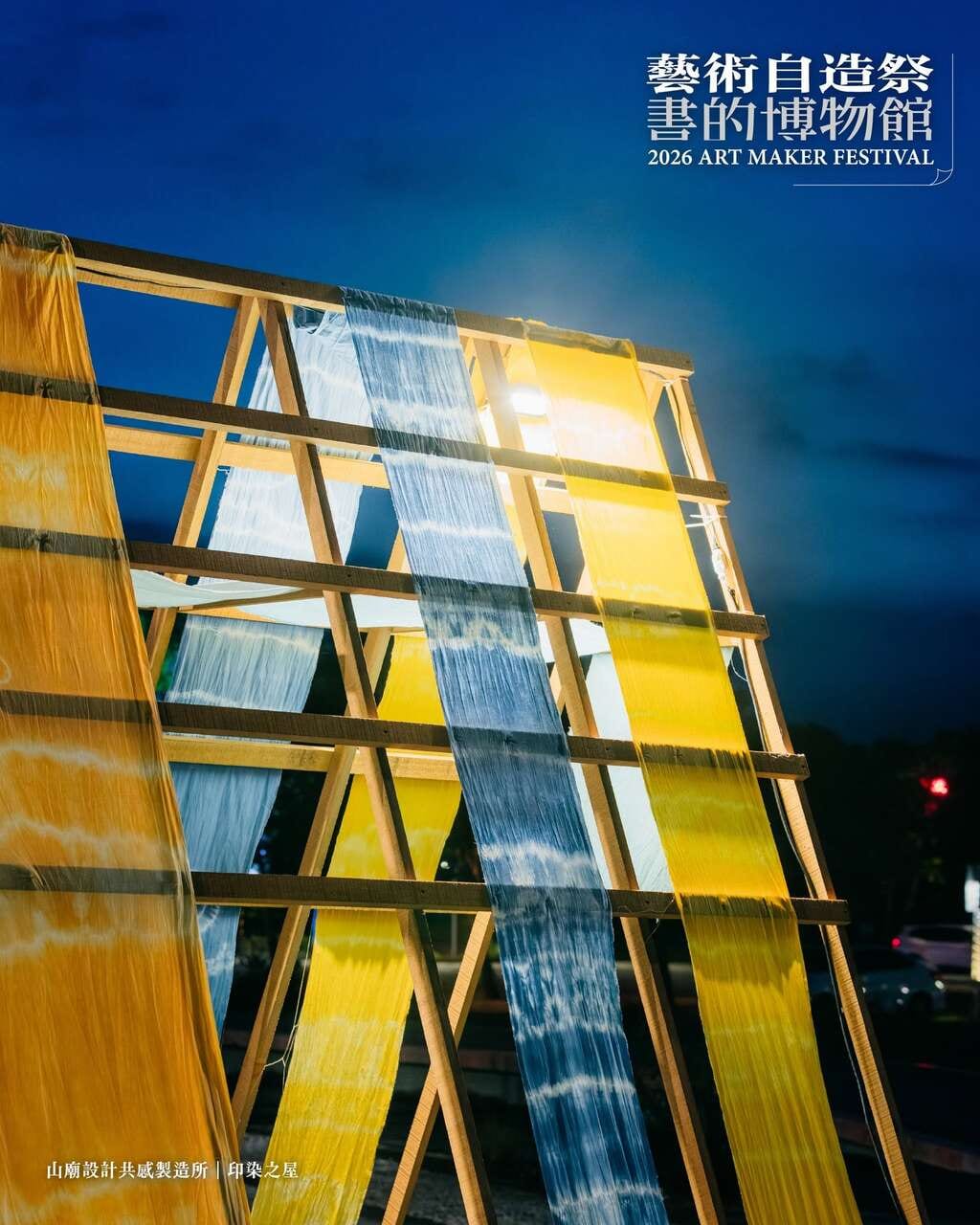 Against a deep blue dusk sky, yellow and indigo dyed cloths hang from a leaning timber frame, with one lamp shining out between them.