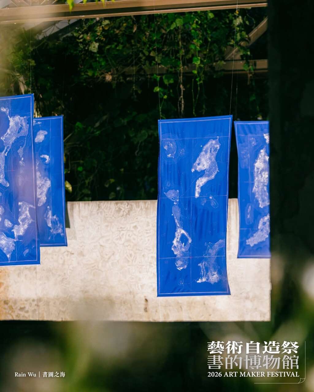 Several ultramarine cloth panels hang under green foliage, curving slightly in the air, each printed with white island-like shapes on a fine grid.