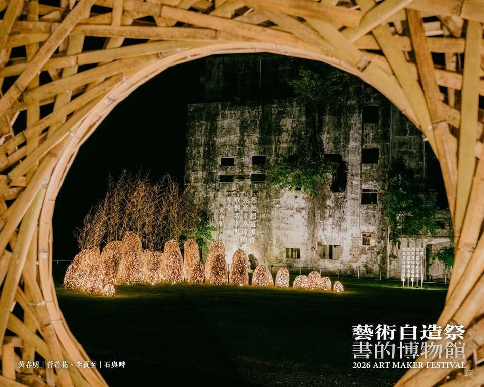 Through a circular bamboo opening, a line of lit stone-like forms stands on night grass before the concrete wall of the former paper mill, with projected text on it.
