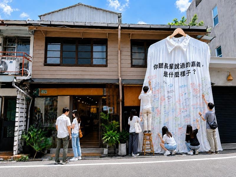 An enormous white shirt hangs across the storeys of an old house; people write on its lower edge.