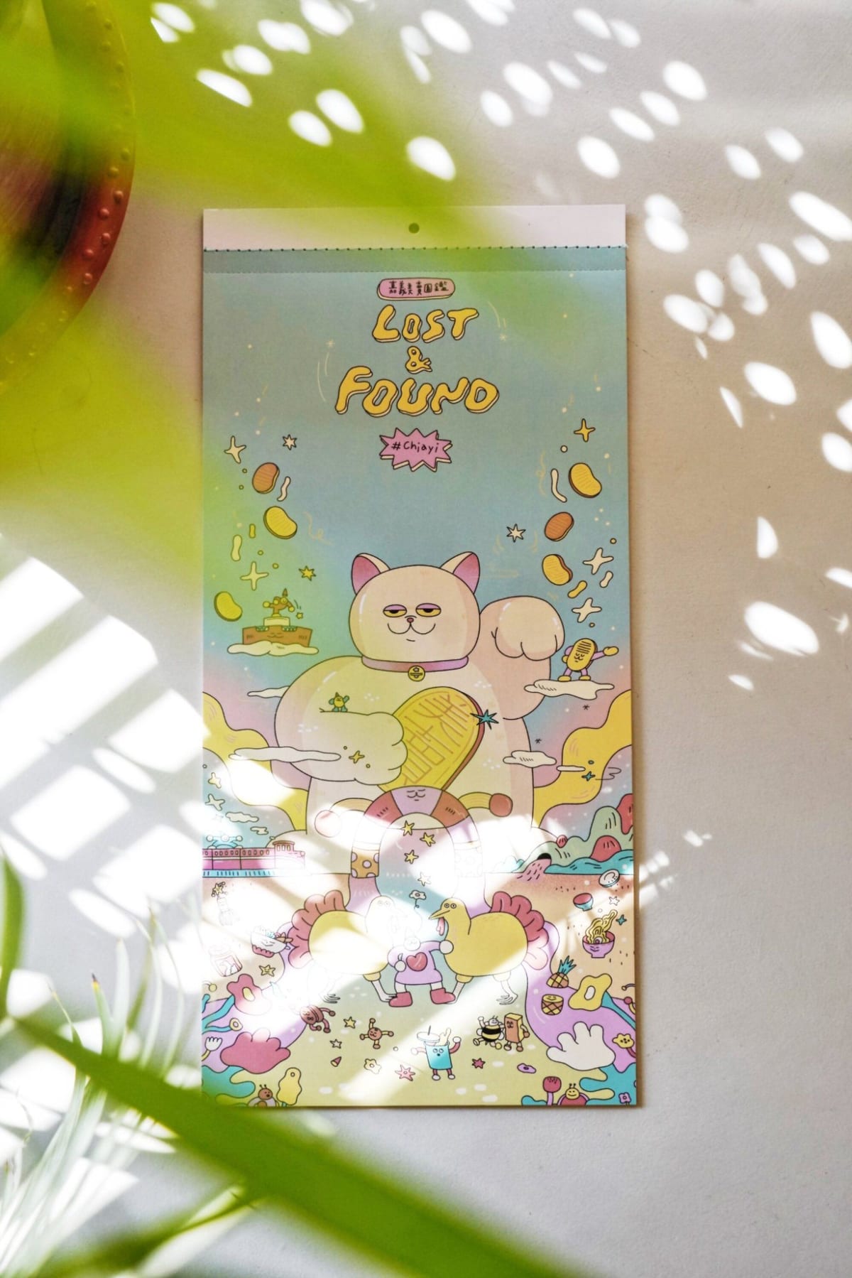 A photographed illustrated Lost & Found calendar with a raised-paw lucky cat, small birds and food characters, under patches of sunlight.