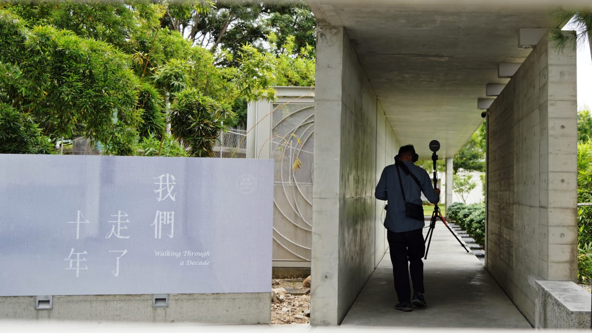 In a concrete corridor a man with a shoulder bag sets up a microphone on a stand facing into the depth of the passage; on the lawn outside stands a sign reading Walking Through a Decade.