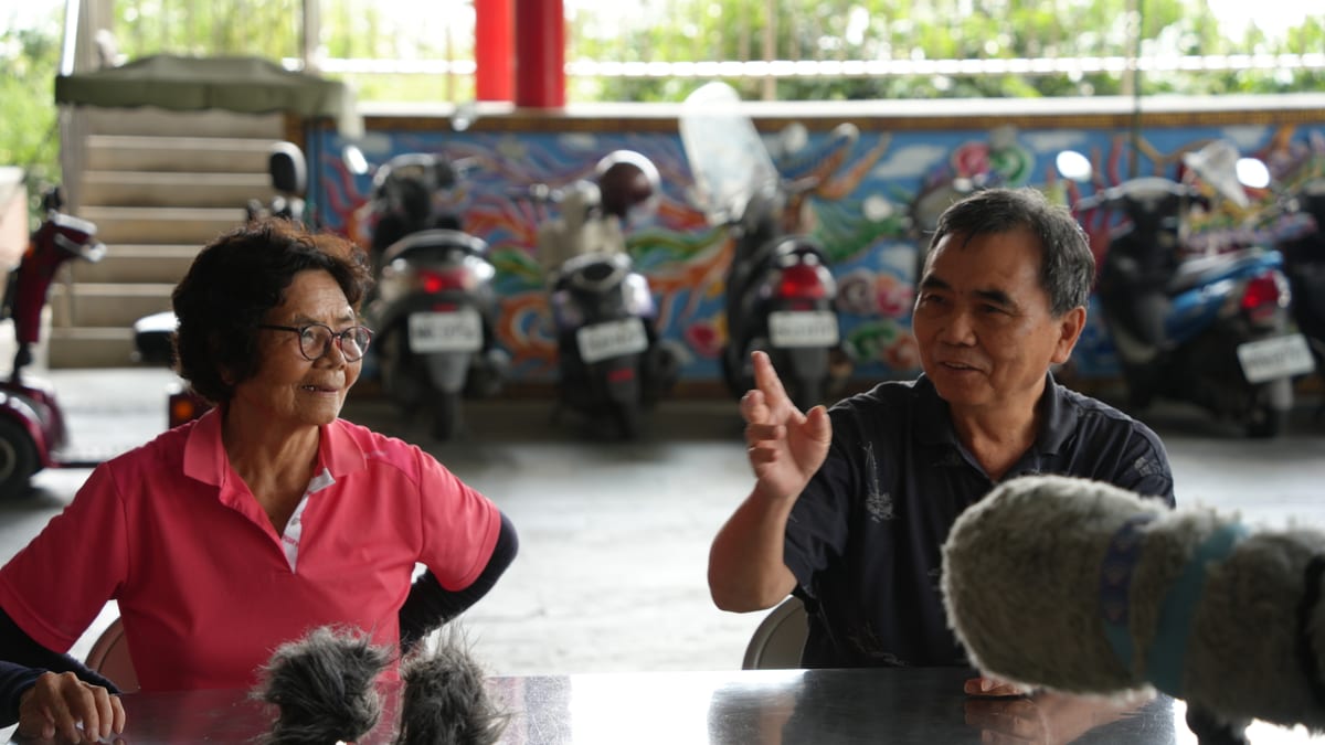 At a metal table under a temple canopy, a woman listens as a man talks with a raised hand; two microphones in furry windshields stand at the near edge of the table, with a row of scooters parked behind.