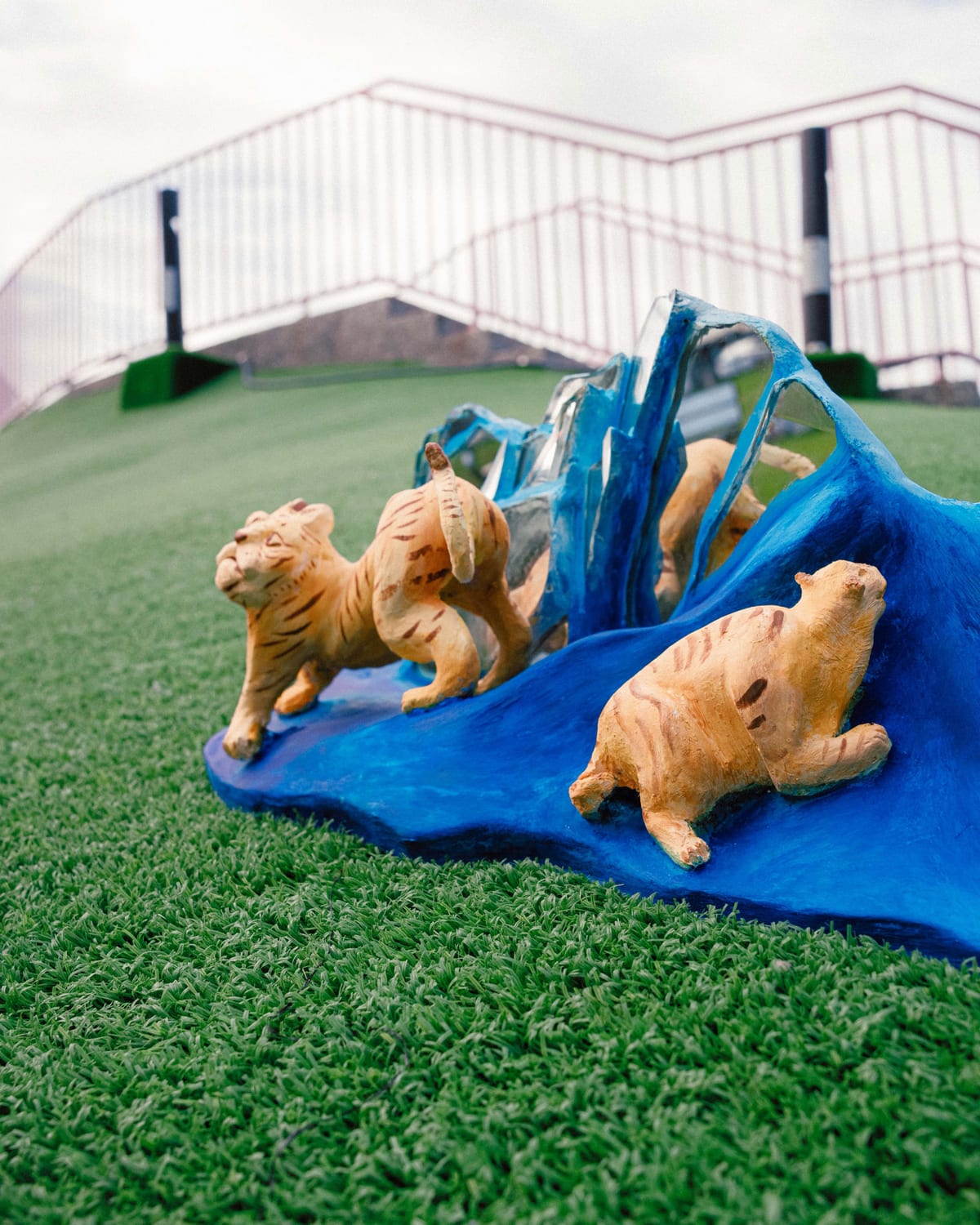 Two ochre clay tigers stand and recline in front of blue mountains with mirrored faces, photographed on green turf.