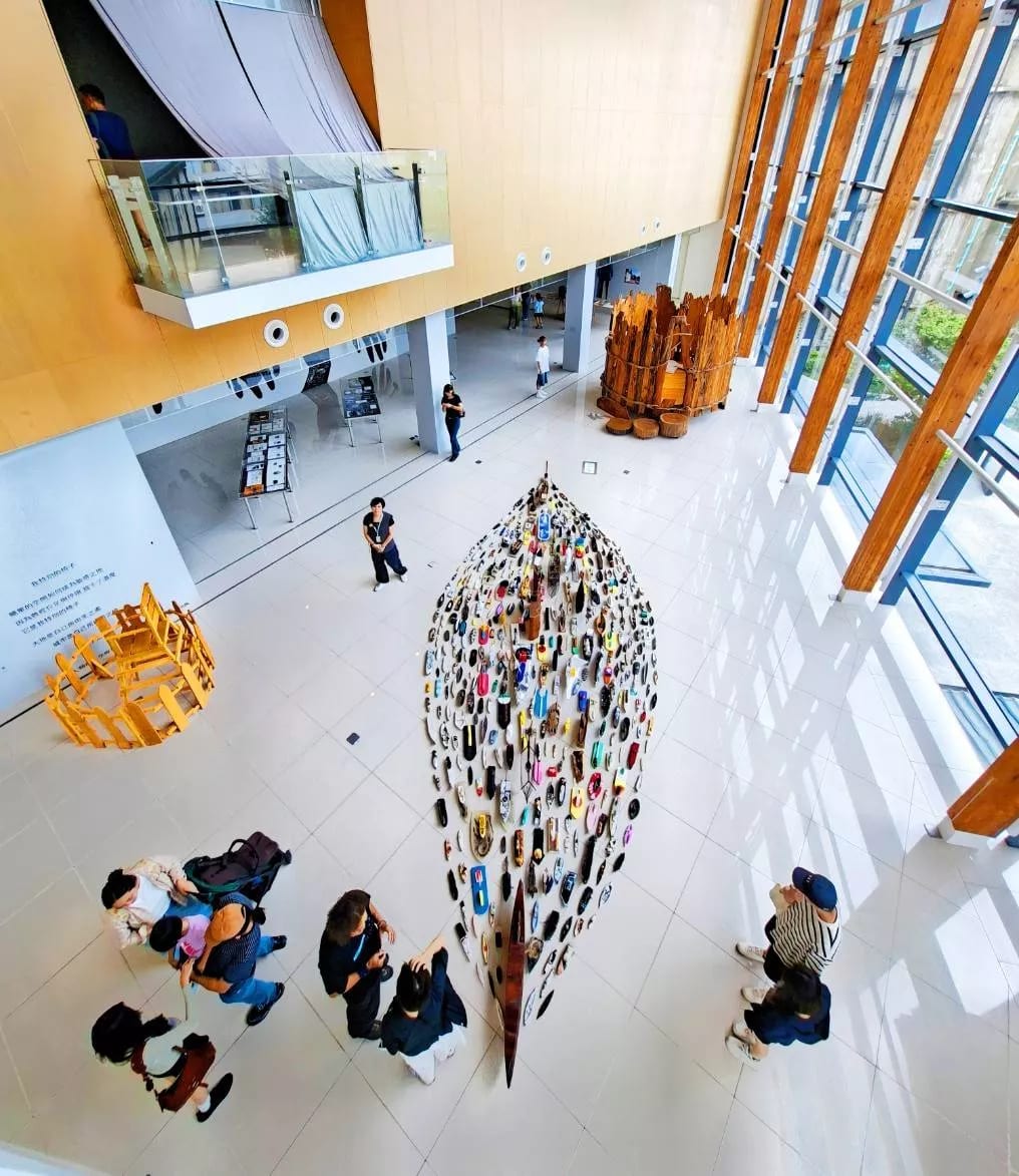 Looking down into the museum's full-height atrium, a long narrow wooden boat rests on the floor hung with rows of small coloured objects, visitors standing around it and a timber-framed glass wall to the right.