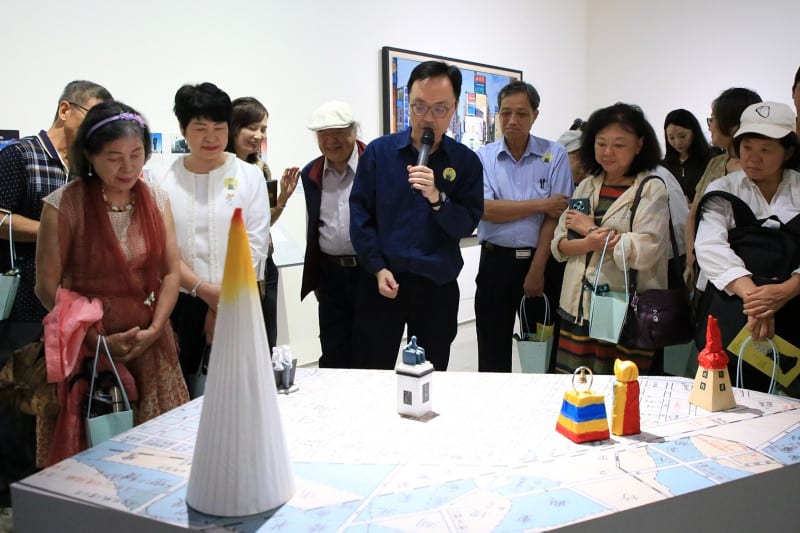 A dozen visitors stand around a long table covered with a hand-drawn street map, on which sit a white cone, small lantern-like houses, and cloth-wrapped statue models; a man at the centre explains into a microphone.