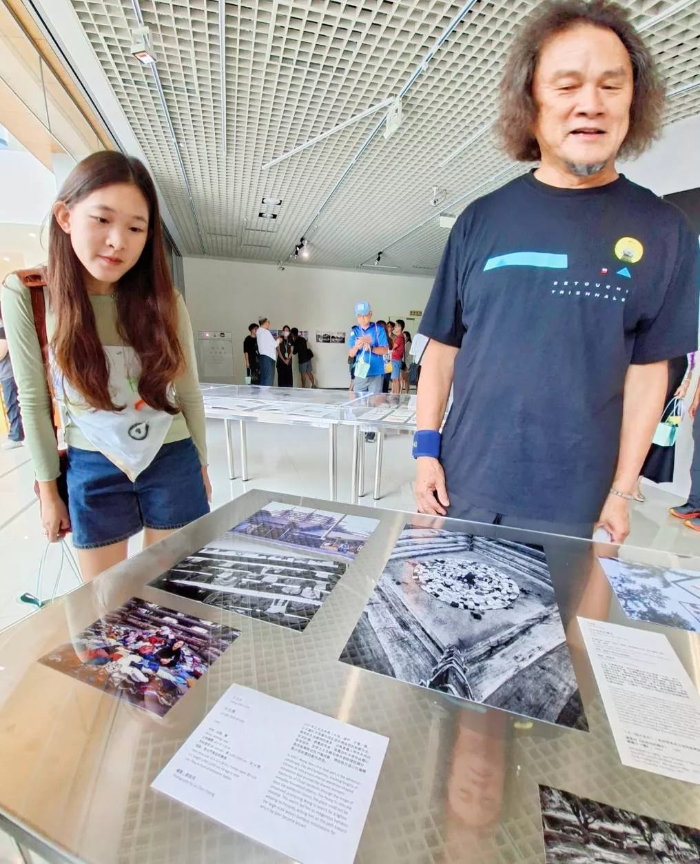 Old black-and-white and colour working photographs lie flat under glass in the gallery; a curly-haired man stands at the right talking while a young woman leans in from the left to look.