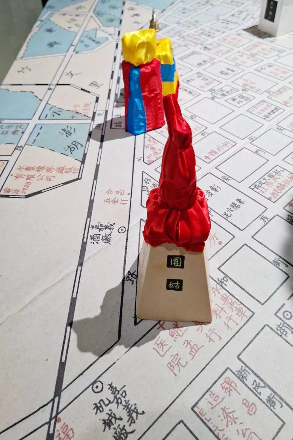A hand-drawn map of Chiayi's streets covers a table, with a small statue wrapped in layers of red, yellow, and blue satin standing on it, the word for unity carved on its base.