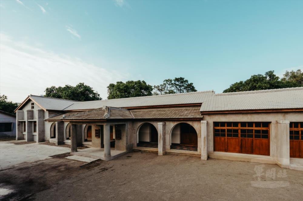 A low grey building with arched openings, timber-framed doors and windows and a tiled roof under a wide blue sky.