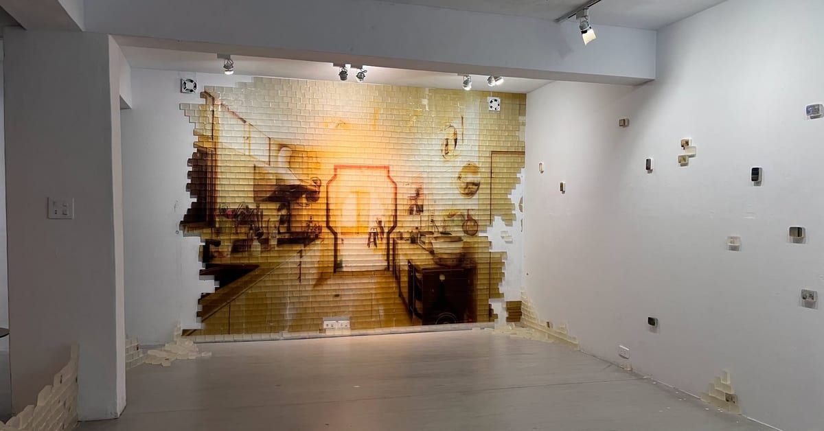 A warm kitchen image covers rows of translucent pale-yellow blocks on a gallery wall. Its uneven edges loosen into pieces at floor level; small picture-bearing blocks dot the adjoining white wall.