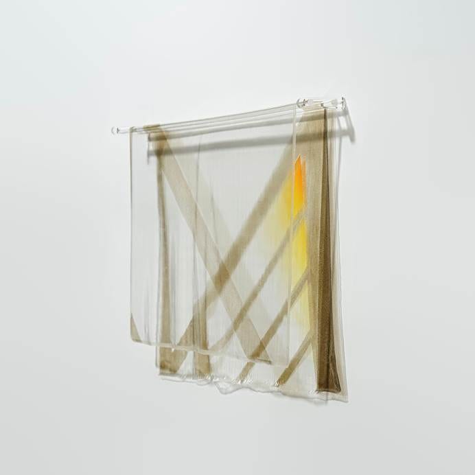 Translucent paint hangs from a clear horizontal rod, with brown diagonal bands, yellow colour and shadows on the white wall.