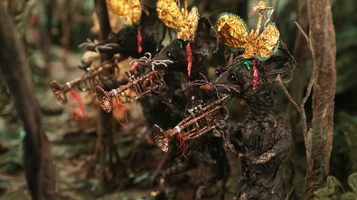 Paper rats wearing elaborate gold headpieces play brass instruments with red threads hanging from them, against a blurred woodland background.