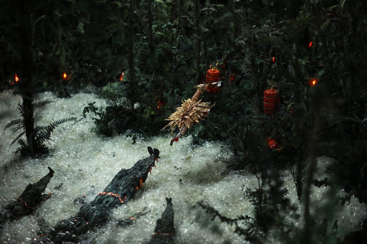 A small paper figure hangs above the open jaws of crocodiles in a dark miniature forest with red lanterns and a pale, foaming river.