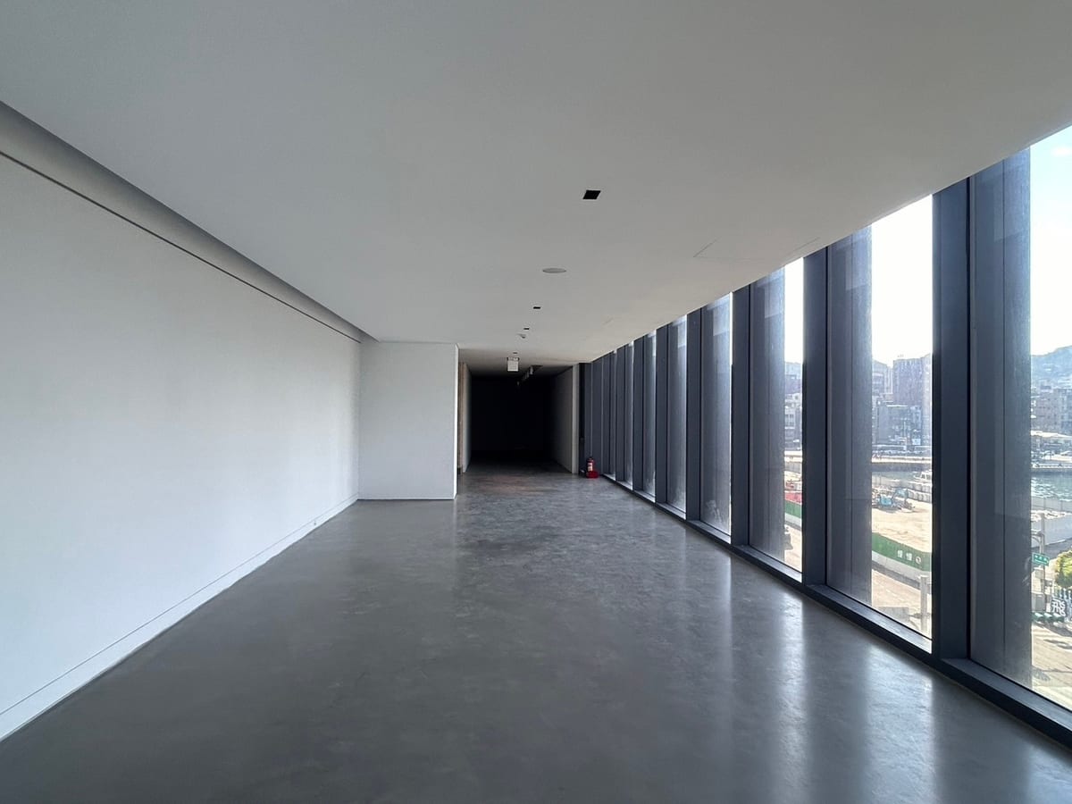 An empty museum room with a long row of floor-to-ceiling windows looking towards the port, daylight across the grey floor.