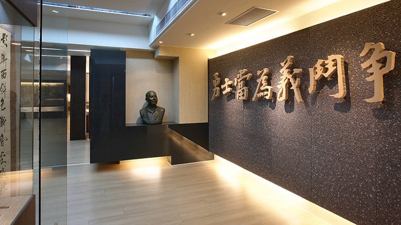 Raised characters reading “a brave man should fight for justice” span a dark museum wall, with a bust of Lai Ho and vitrines to the left.