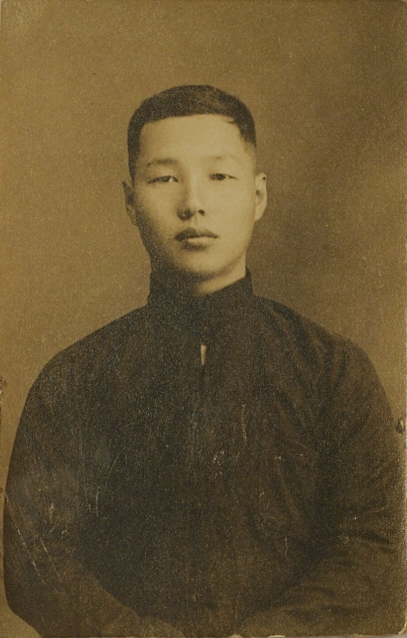 A young Lai Ho sits facing the camera in a Taiwanese-style jacket against a plain studio backdrop.