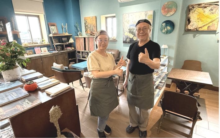Cao Kai-zhi and Li Ruo-mei stand inside Little Island Universe among wooden furniture, books, paintings and ceramics.