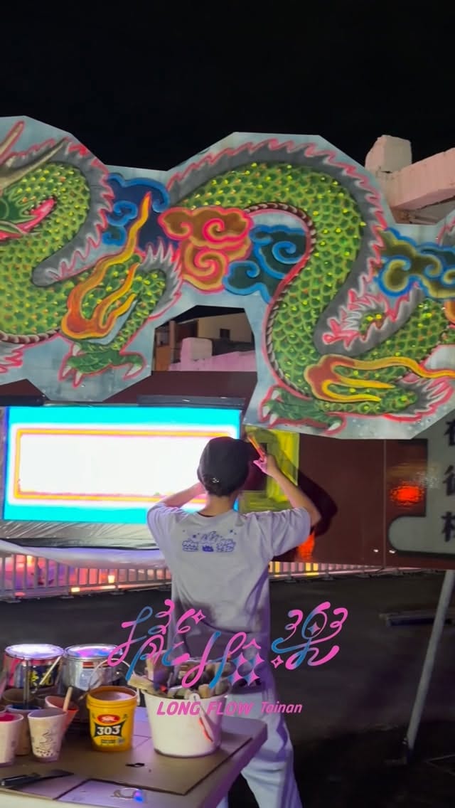 At night on the Kangle Street side, a painter works high on a large dragon form, its green scales edged in fluorescent line; paint cups, a folding table and a light box stand on the ground below.