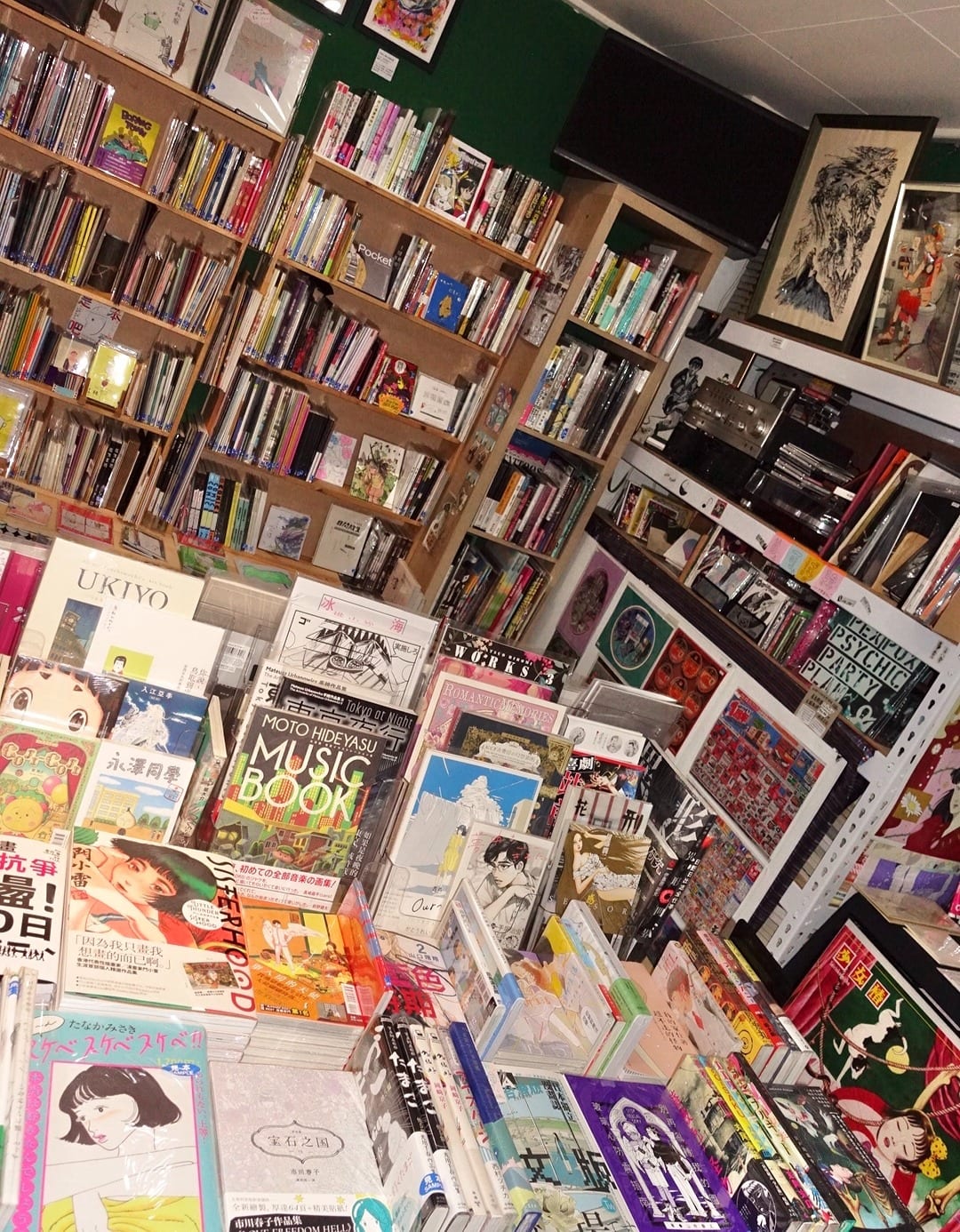 A tilted photograph shows rows of comics and art books on wooden shelves and tables, with framed pictures against a dark-green wall.