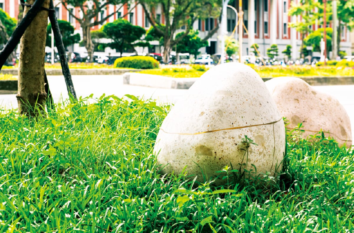 A pale terrazzo sculpture from Chiu Cheng-Hung's Embroidered Swallows # NTUH sits in grass, a fine brass seam running around its rounded surface, with trees and the hospital streetscape behind it.