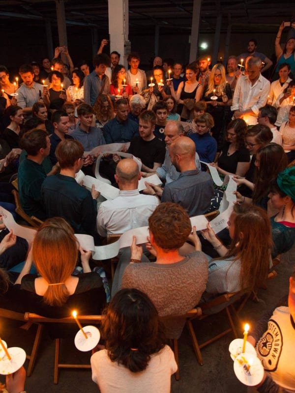 People seated in close circles pass and read a long pale strip; a surrounding crowd holds candles.