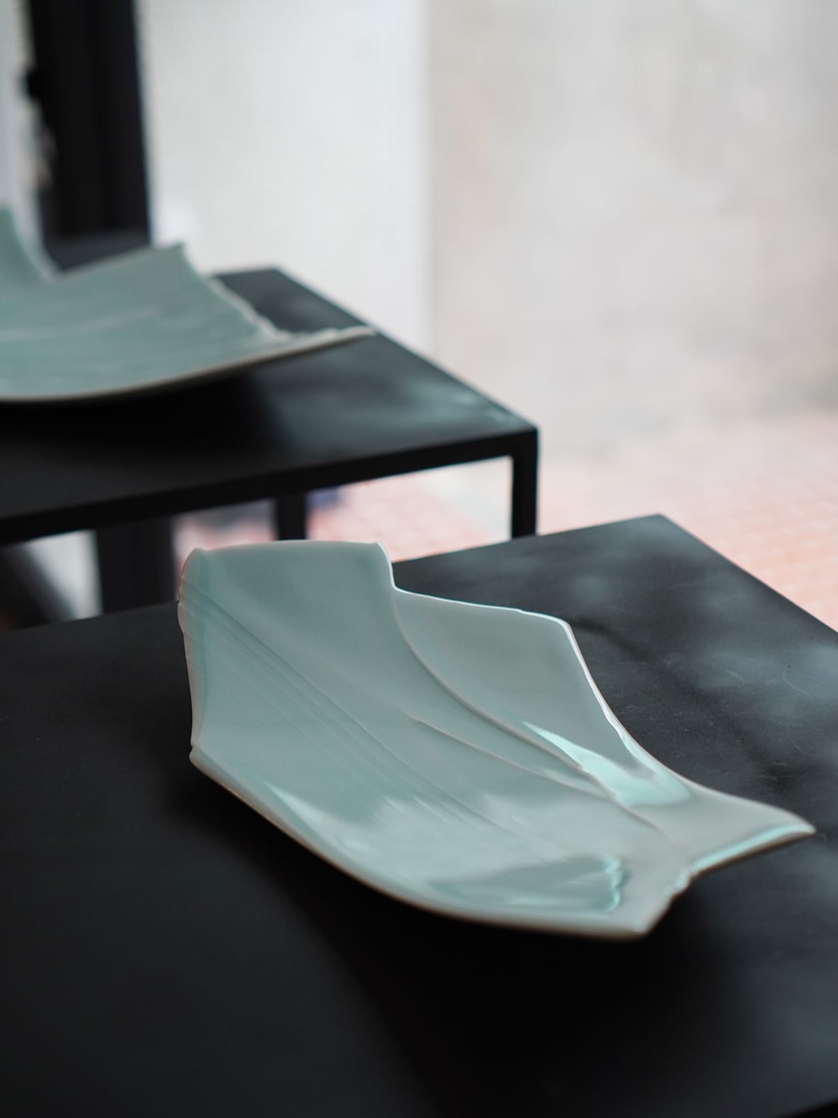 Two pale celadon dishes sit on black plinths, one in front and one behind; each is folded and ridged like creased paper with an uneven edge, the further one out of focus against a grey wall.