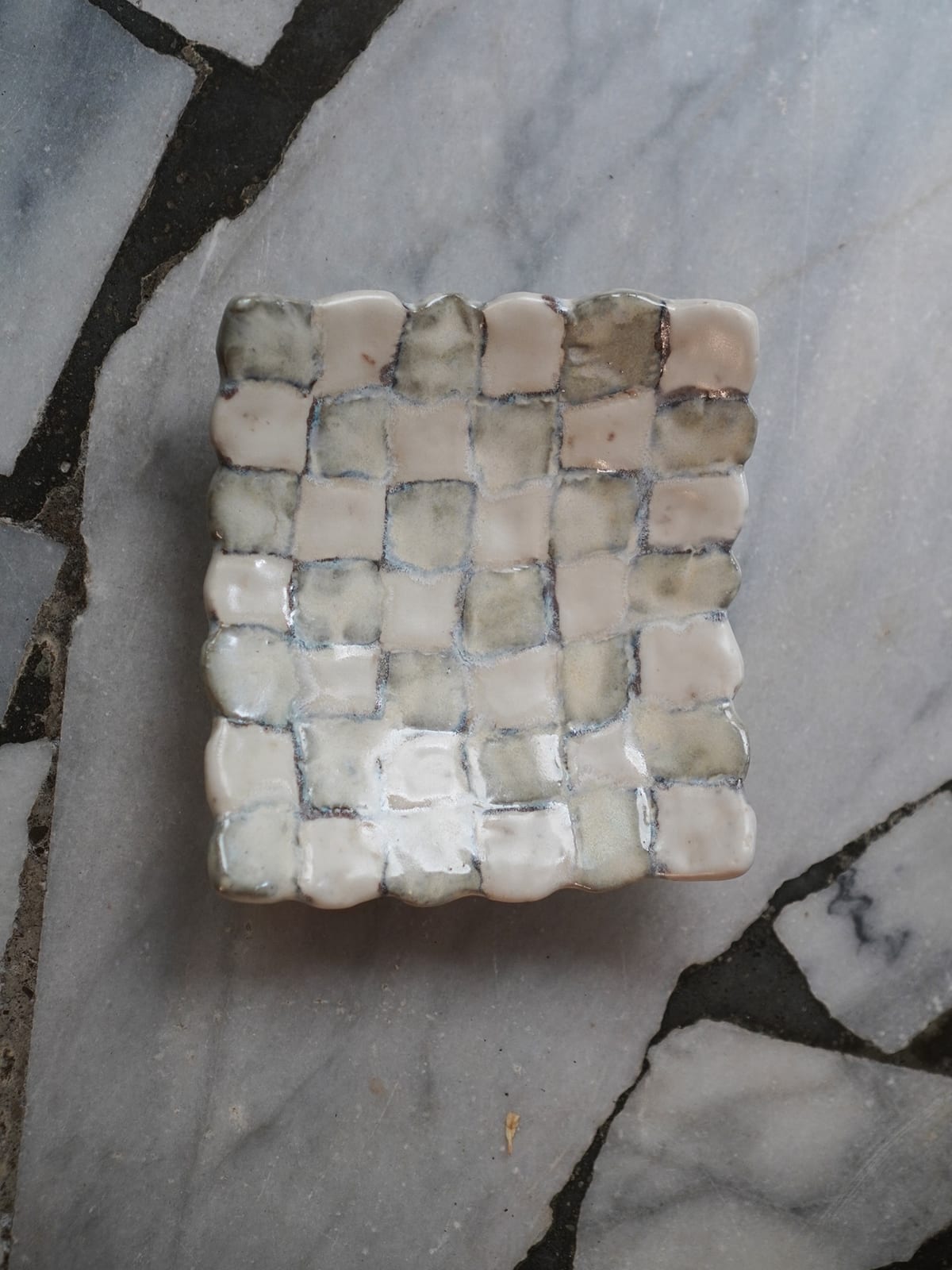 A shallow square dish rests on pale grey marble paving; its surface is a chequerboard of cream and soft grey-green squares with sunken grid lines and a wavy rim.