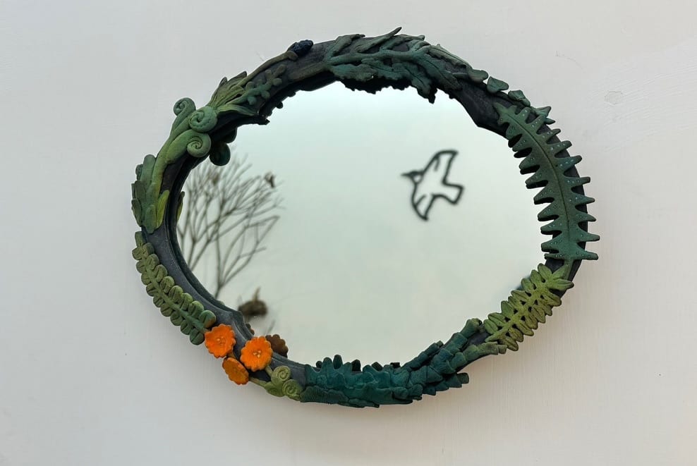 An oval mirror hangs on a white wall inside a ceramic frame modelled as ferns, curling tendrils, and small orange flowers in deep green and blue-green; the glass reflects a bare tree and carries the outline of a black flying bird.