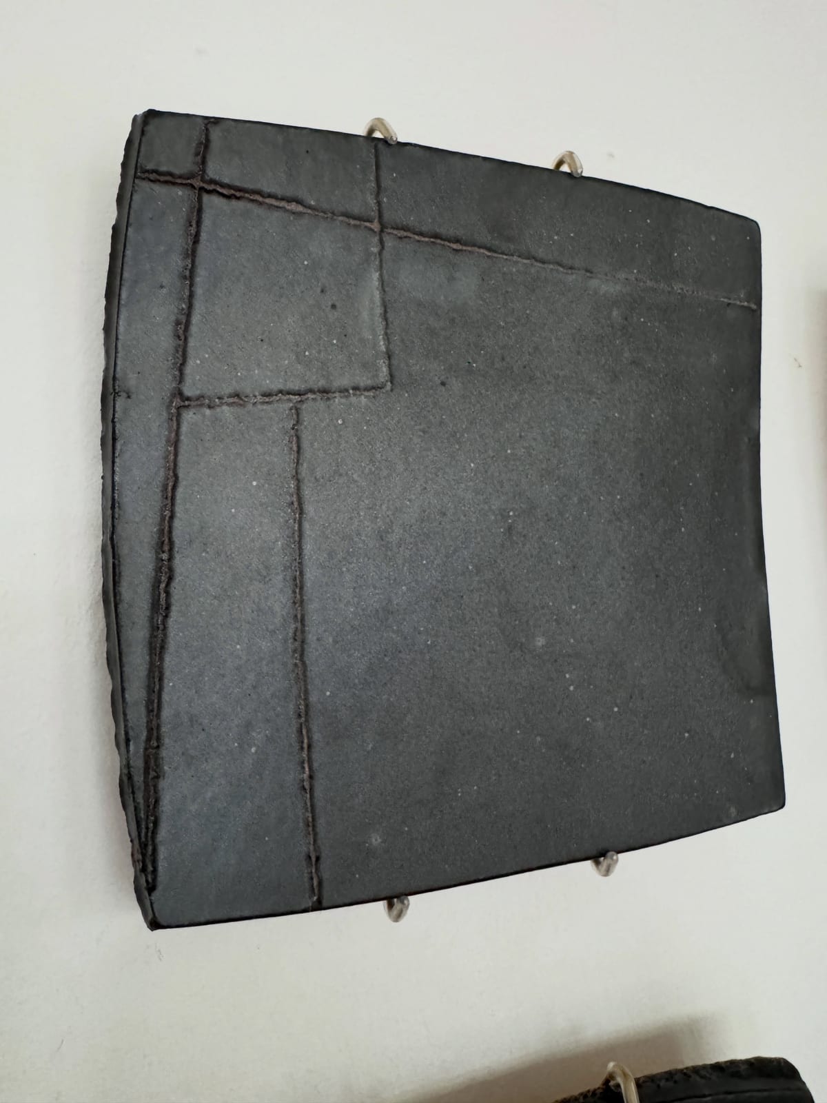 A close view of one black square ceramic panel held to a white wall by metal hooks; incised lines cross the matte surface, dividing it into unequal rectangles.