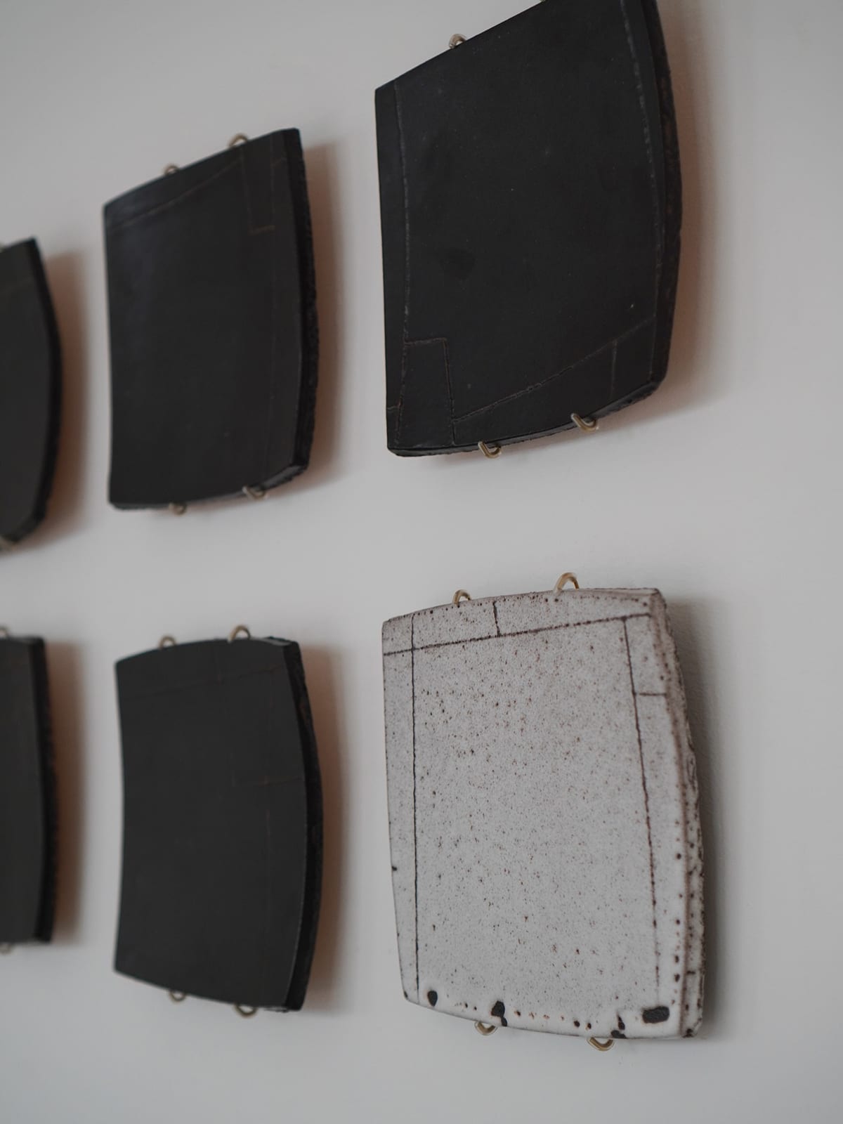 Several slightly curved square ceramic panels hang on a white wall from small brass hooks; most are matte black, one is a speckled white glaze, each incised with fine straight dividing lines.