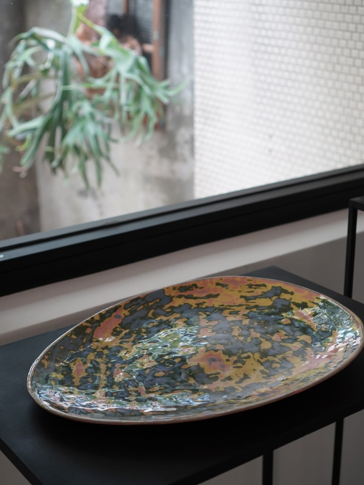 A large oval platter sits on a black metal stand by a window, its glaze pooling in yellow, green, blue, and ochre; outside, a staghorn fern hangs against a tiled wall.