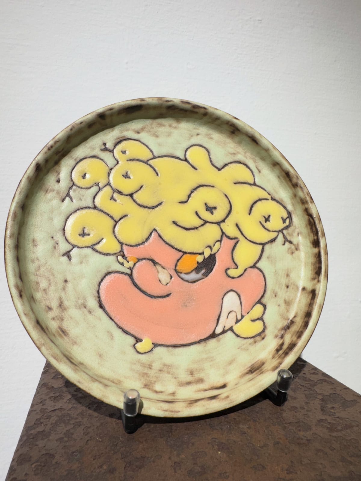 A pale green round plate stands on a display easel, painted with a plump pink-orange figure carrying a cluster of yellow fruit-like forms above its head; brown speckles ring the rim.