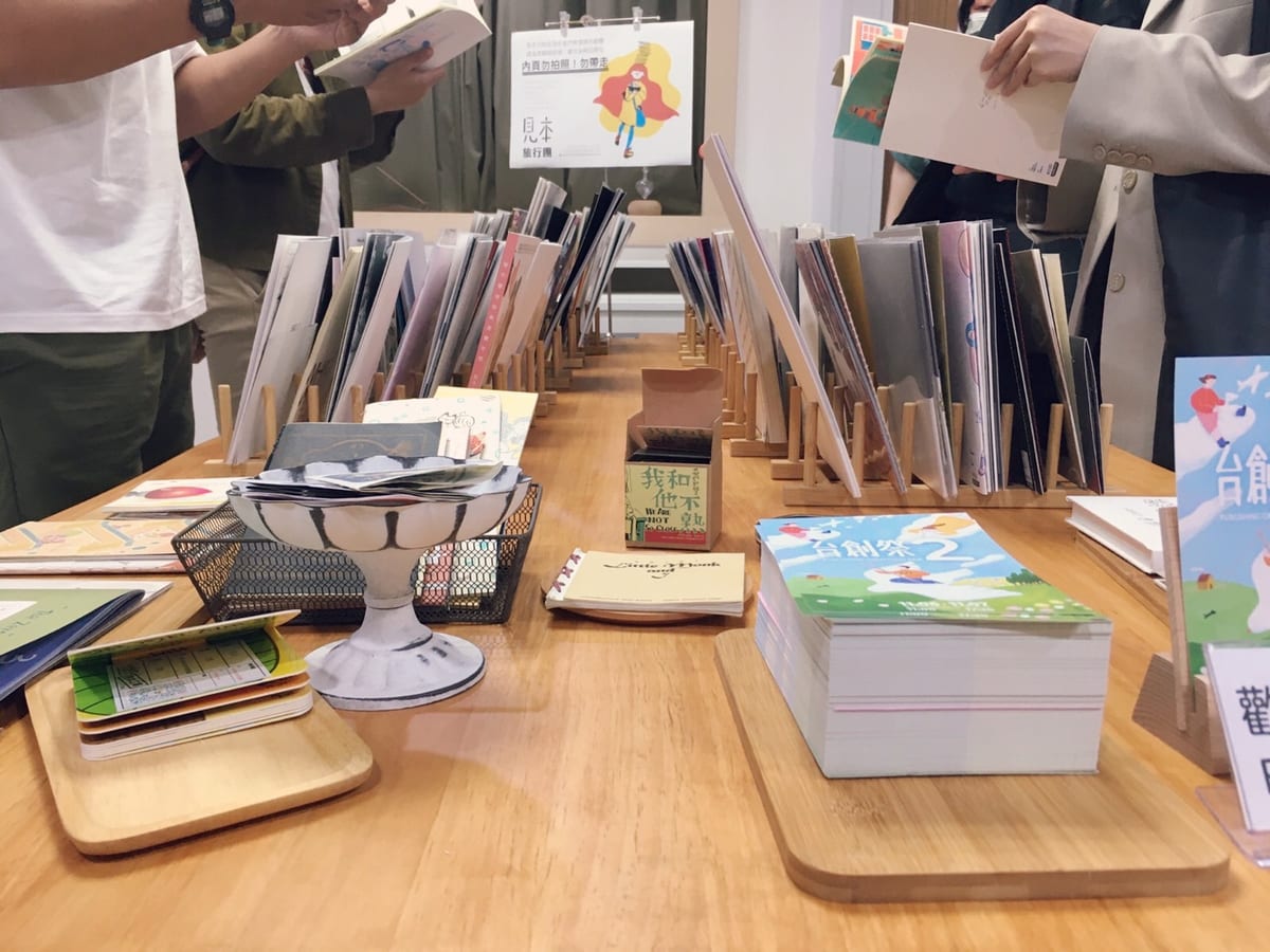 An archival organiser photograph of sample reading: zines and illustrated books stand in wooden racks while visitors browse them.