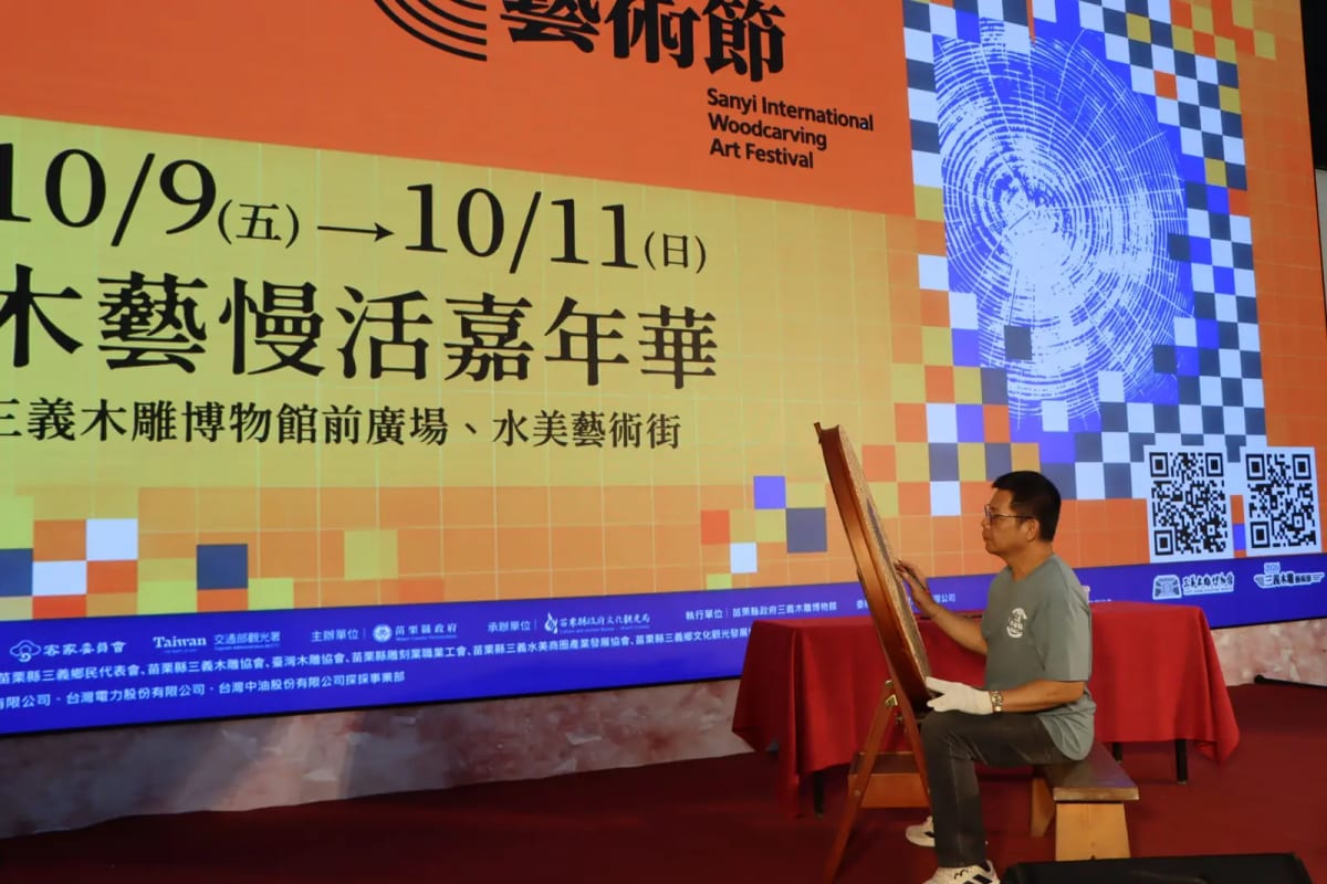 A seated craftsperson carves an upright wooden panel in front of the festival backdrop.