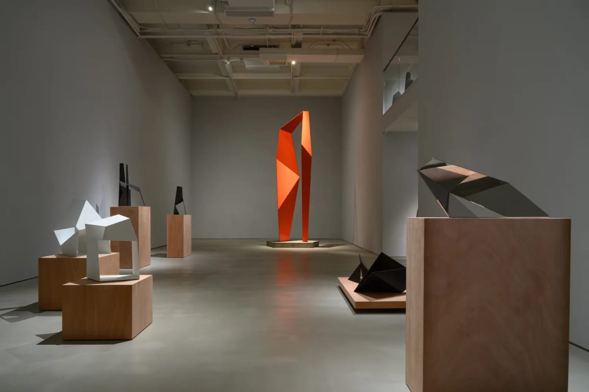 A white gallery where small metal sculptures sit on wooden plinths and low platforms, with a tall orange-red work standing against the far wall.