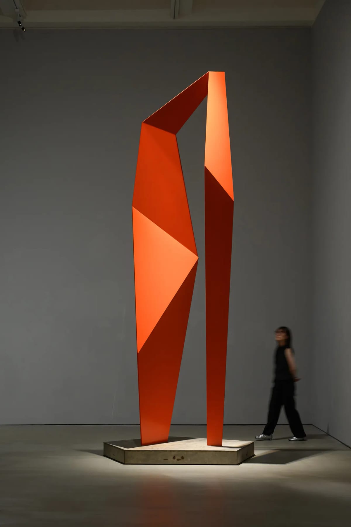 Two long orange-red legs rise from a low base and meet through folded planes around a narrow central opening, with a person small beside the work.