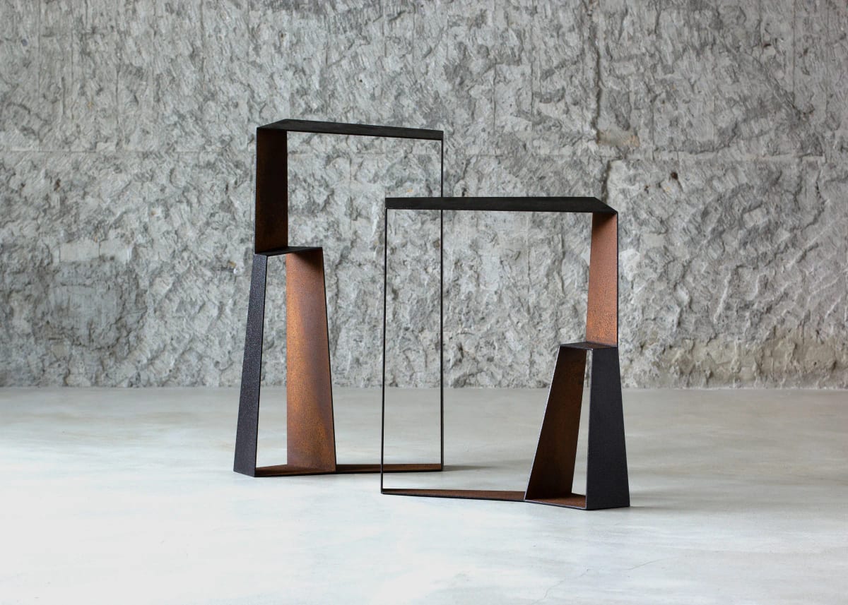 Two upright iron units stand side by side, each with a bent perimeter, narrow rods, and a rectangular opening; one outline overlaps the other and the ground stays visible through both.