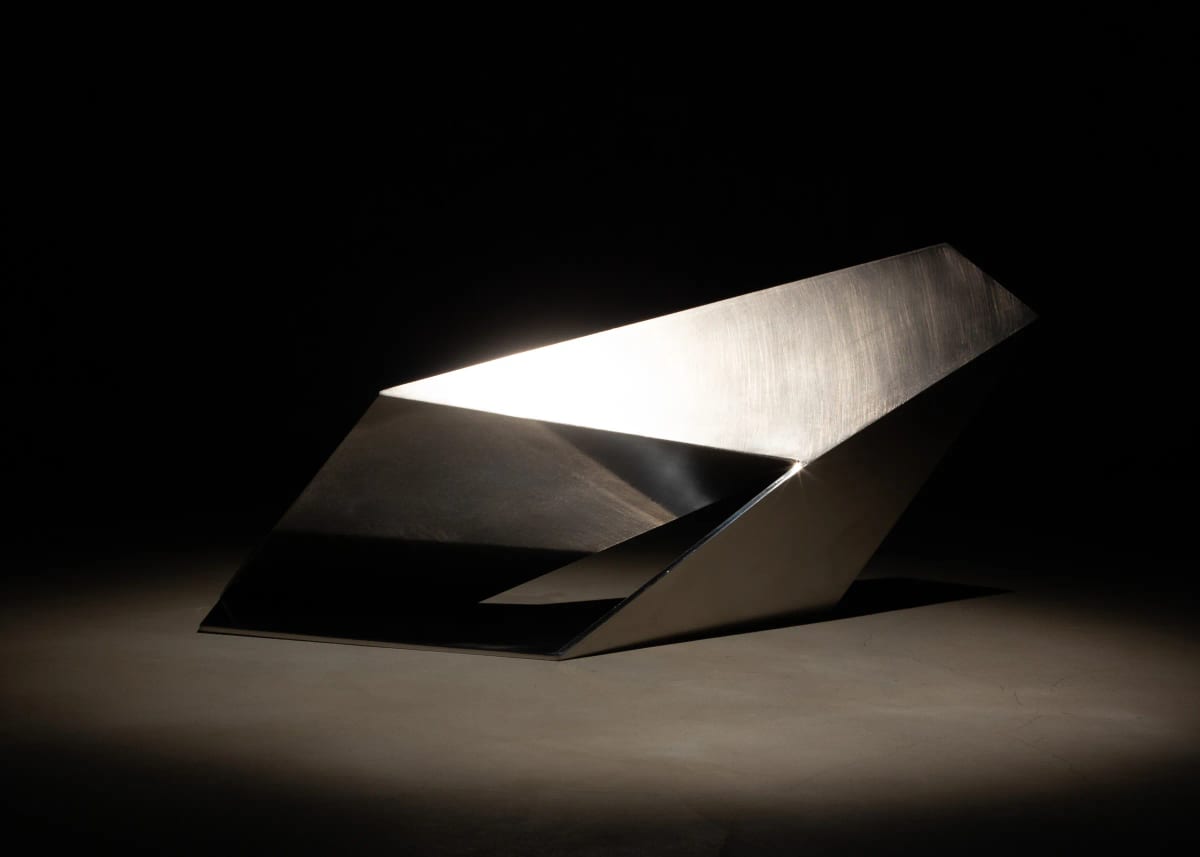 A low stainless-steel form of folded planes leaves an opening at its centre, a bright band along one upper plane and darker reflections below.
