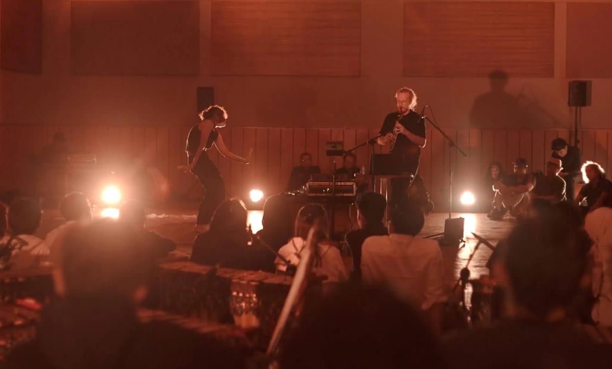 Under warm amber lights, a dancer bends forward beside a clarinet player. Listeners sit around them, with electronic equipment behind the performers.