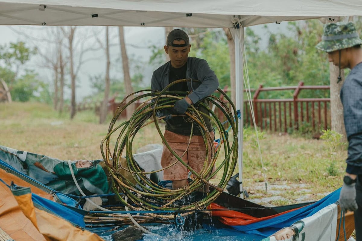 Under a white canopy a man in a cap lifts a woven bamboo sphere dripping out of a water tank, another worker standing to his right.