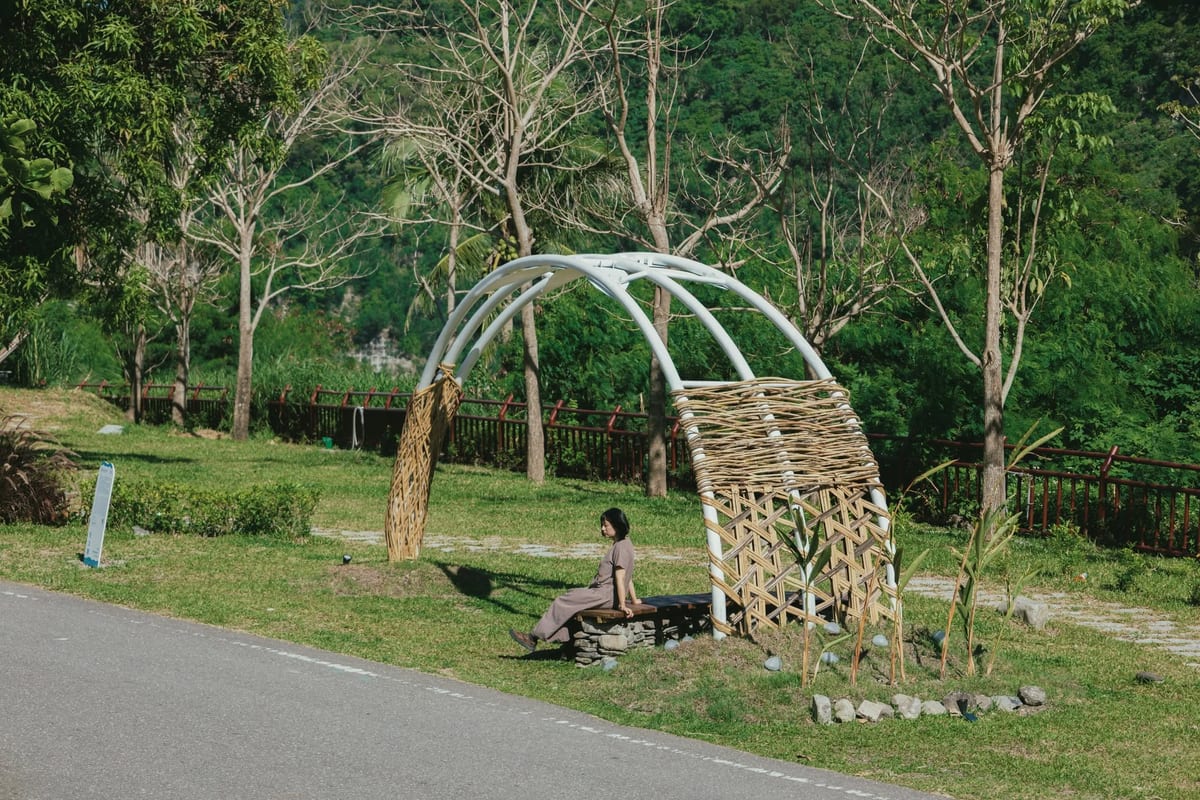 On riverside grass, white steel arches cross into a half-open shelter with one side wrapped in woven bamboo; a woman sits on a stone bench beneath it, with the valley and forest behind.
