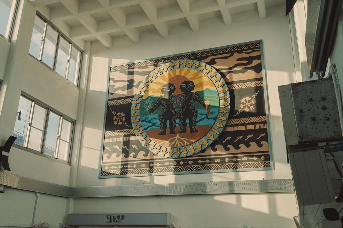A large rectangular woven panel hangs on the white wall of a high station hall, its centre a circle of sunrise and two figures inside geometric borders, with a sign reading Tickets below.